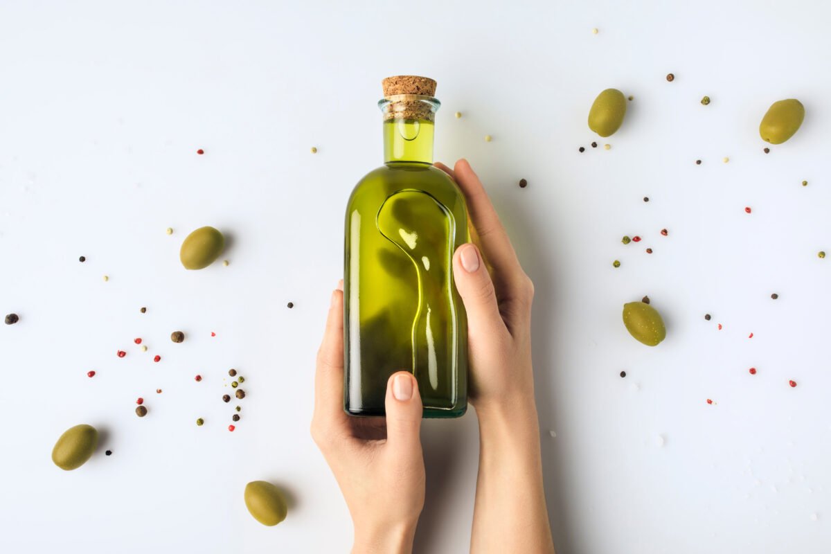 A pair of hands holding a bottle of olive oil, surrounded by green olives and peppercorns on a white background.