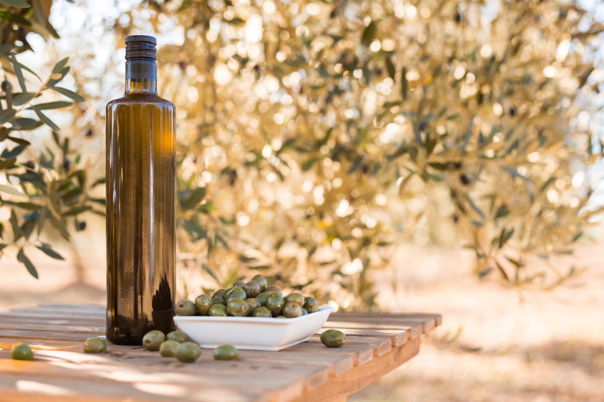 A bottle of olive oil with a bowl of green olives on a wooden table, set against an olive tree background.